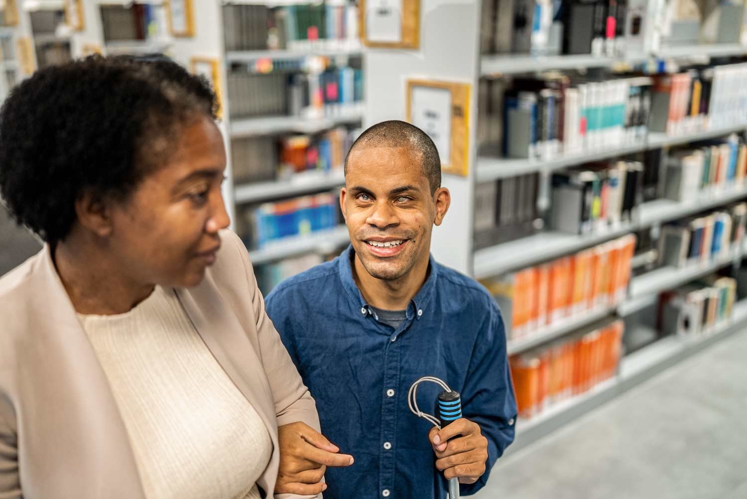 young man of color being led through a library while holding a white cane