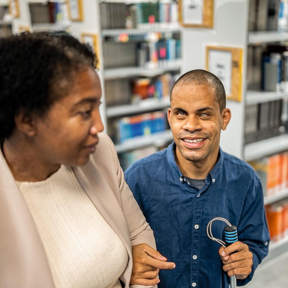 woman leading a man holding a white cane through a library